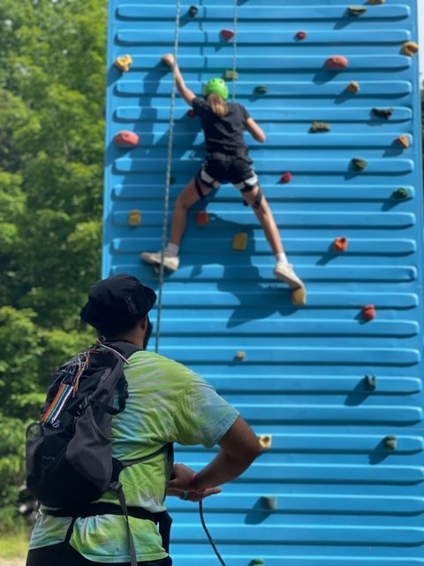 Child wearing a green helmet and black shorts climbing a blue outdoor rock wall, with a person in a tie-dye shirt and black cap spotting from below.