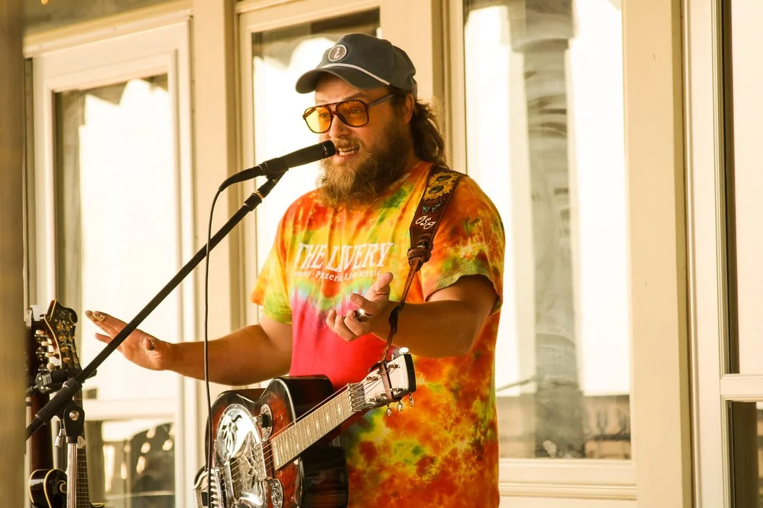 A man with long hair and a beard, wearing sunglasses, a tie-dye shirt, a cap, and playing an acoustic guitar while singing into a microphone.