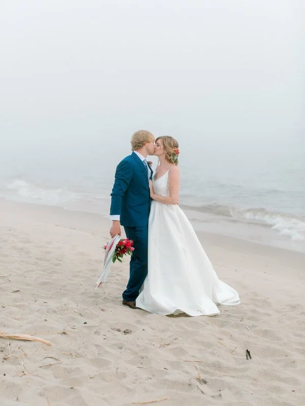 A bride and groom kissing on a foggy beach, with the bride in a white wedding dress and the groom in a blue suit holding a bouquet of flowers.