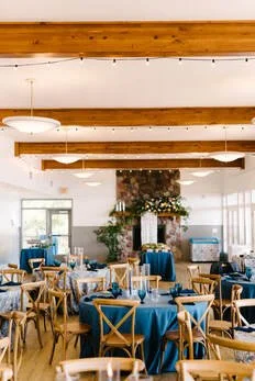 Elegant event space with round tables covered in blue tablecloths, wooden chairs, floral centerpieces, a white fireplace, and large windows letting in natural light.