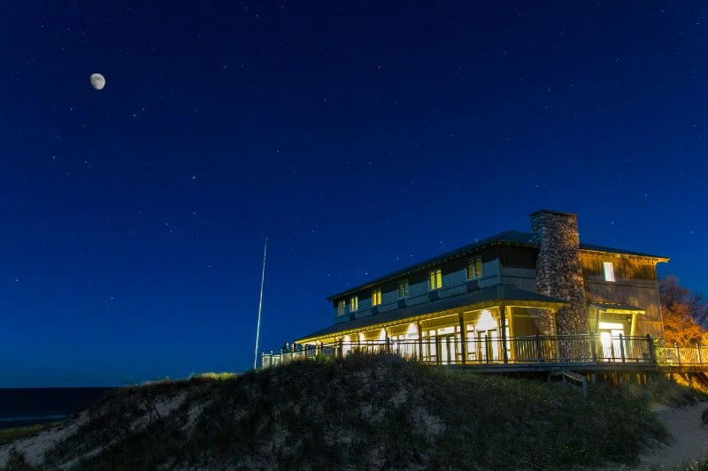 A house on a hill at night with lights on, sky filled with stars and the moon visible.