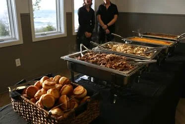 Buffet table with trays of cooked meats and a basket of bread rolls in the foreground, two people standing behind the table.