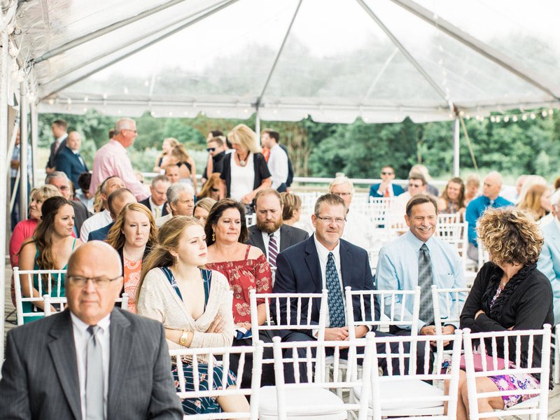 People attending a formal outdoor event inside a large white tent, sitting on white chairs and dressed in business or semi-formal attire.