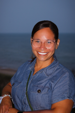 Woman smiling outdoors near the ocean at dusk wearing a denim shirt and glasses