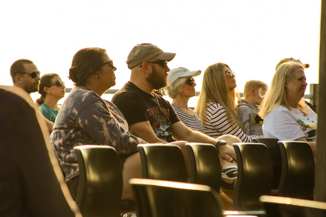 Group of people sitting outdoors, wearing sunglasses and casual clothing, watching an event during sunset.