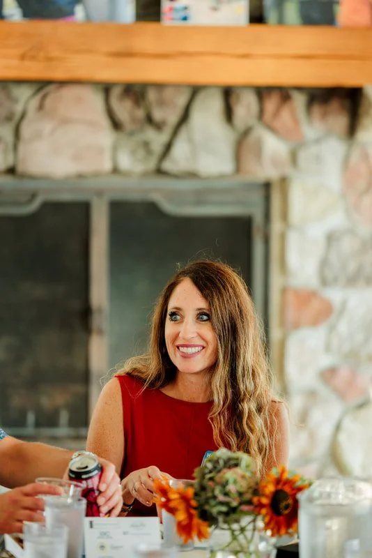 A woman with long, wavy brown hair, wearing a sleeveless red dress, sitting at a table with a floral centerpiece, smiling and looking to her right inside a cozy room with a stone fireplace.