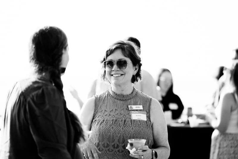 Two women talking at a social event, one wearing sunglasses and holding a drink, smiling.