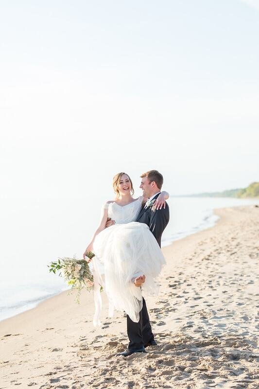 A newlywed couple on the beach, with the groom carrying the bride and both smiling, the bride holding a bouquet of flowers.