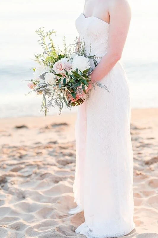 A bride in a white strapless wedding dress holding a bouquet of pink and white flowers with greenery, standing barefoot on a sandy beach with the ocean in the background during sunrise or sunset.