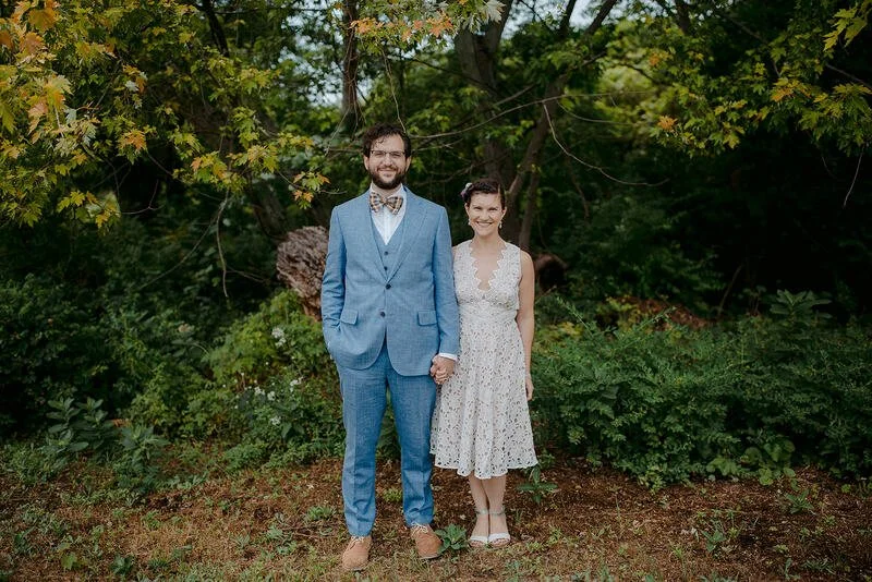 A man in a blue suit and a woman in a white dress stand hand in hand in an outdoor setting with green trees and bushes.
