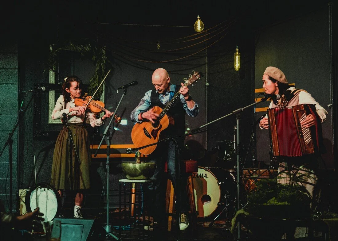 Three musicians performing on stage: a girl playing a violin, a man playing an acoustic guitar, and a woman playing an accordion.