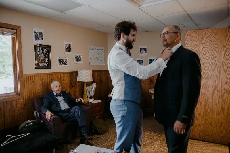 Three men in a room with wooden paneling, two are standing close face-to-face, one fixing the other's collar; the third man is sitting on a couch observing. The room has framed photos and posters on the wall, a window, and a lamp.