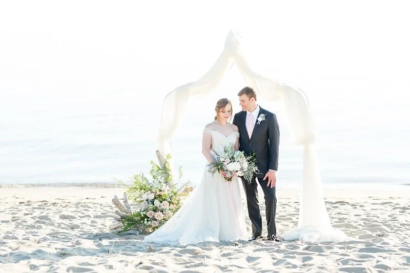 A bride and groom stand under a white wedding arch on a beach, with the bride holding a bouquet of flowers, during their wedding ceremony.