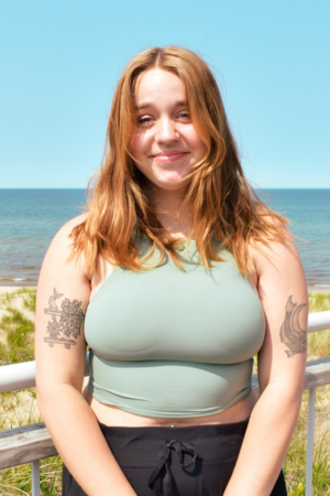 Young woman with red hair standing outdoors at the beach, smiling, wearing a sleeveless light green top and black shorts.