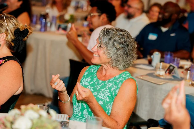 An older woman with gray curly hair wearing a green sleeveless dress with white floral patterns, smiling and gesturing with her hands at a gathering or celebration.