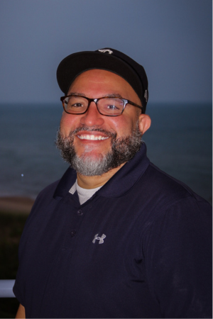 A man with glasses and a beard smiling outdoors at the beach, wearing a black cap and a dark Under Armour jacket.