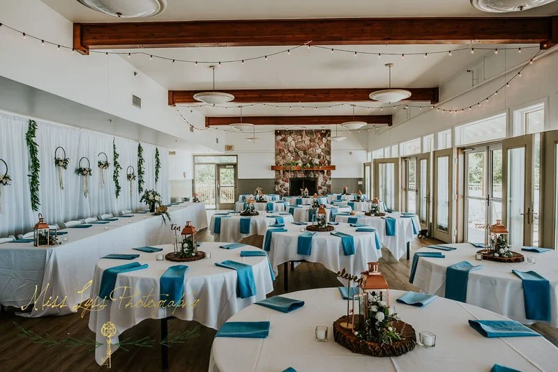 A banquet hall decorated for a formal event with white tablecloths, blue napkins, and centerpieces, featuring a stone fireplace and large windows letting in natural light.