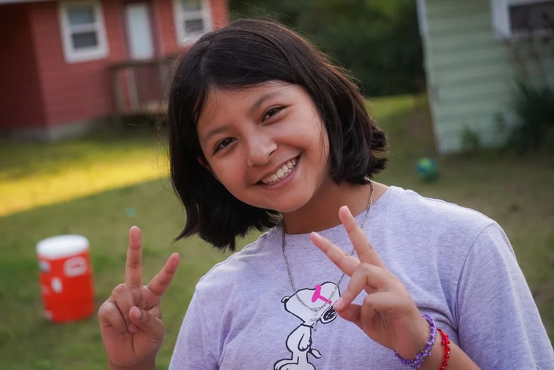 A young girl with short dark hair smiling outdoors, making peace signs with both hands