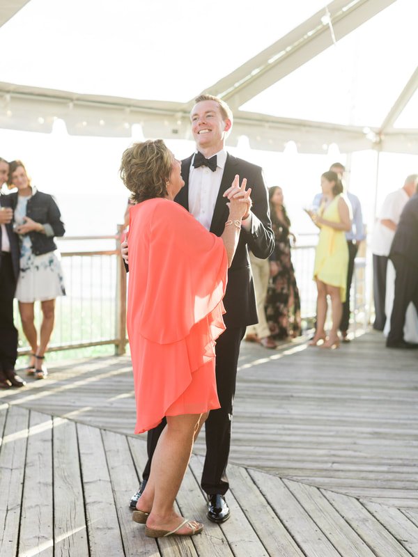 A young man in a black tuxedo dancing with an older woman in a coral pink dress at an outdoor event, with other guests in the background.