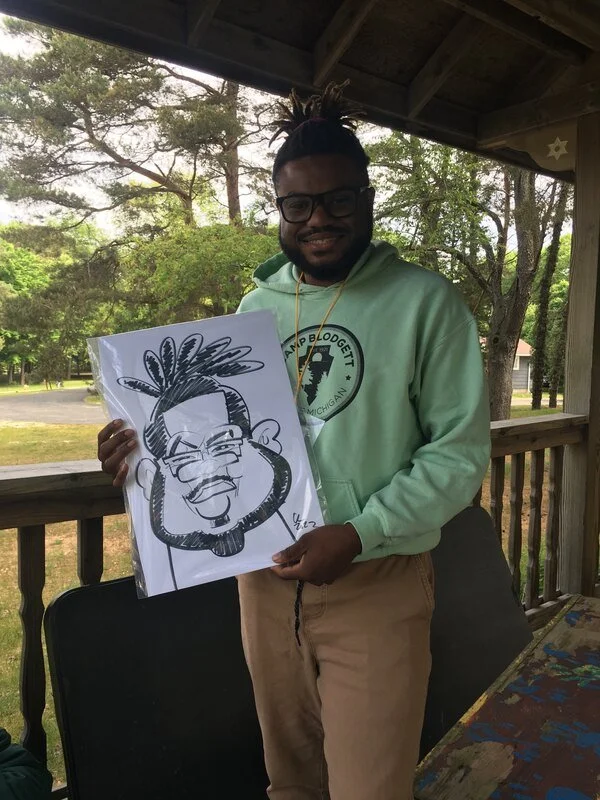 A smiling man with glasses and dreadlocks holding a caricature drawing of himself, standing on a porch surrounded by trees.