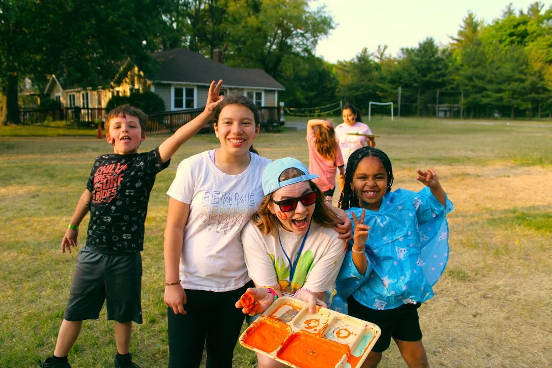 Group of children and a young woman outdoors on a grassy field, smiling and posing with a tray of face paint, with trees and houses in the background.