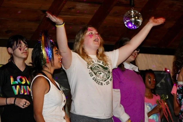 A group of young women at a party dancing under a disco ball, with colorful makeup and festive clothing.