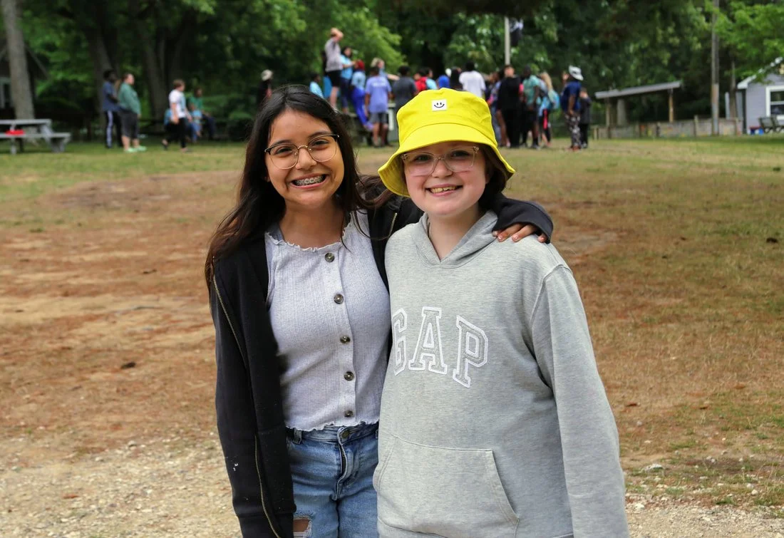 Two young girls smiling and posing outdoors, one wearing glasses, a grey hoodie with "GAP" on it and a yellow hat, the other wearing glasses and a light jacket, with a group of people in the background.