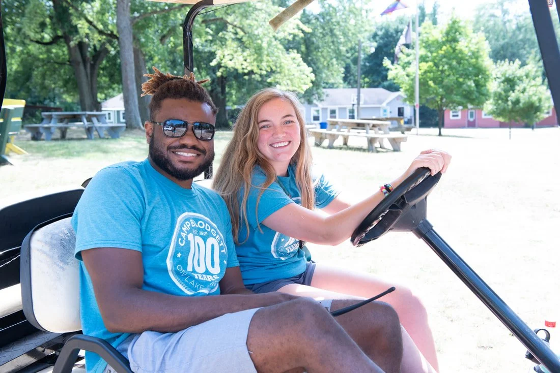 A smiling man and woman sitting in a golf cart outdoors at a camp, with trees and picnic tables in the background.