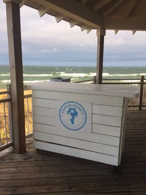 A wooden beachside kiosk with a Camp Blodgett logo, situated on a deck overlooking the ocean with waves and a cloudy sky.