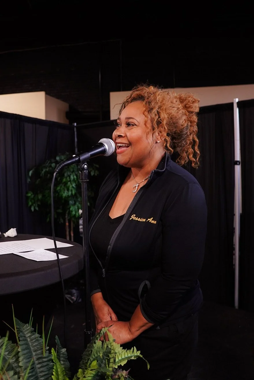 A woman with curly, blonde hair speaking or singing into a microphone at an event, wearing a black jacket with gold embroidery that says "Jessica Ann." She is smiling and standing beside a music stand or table with papers, in a room with black curtai