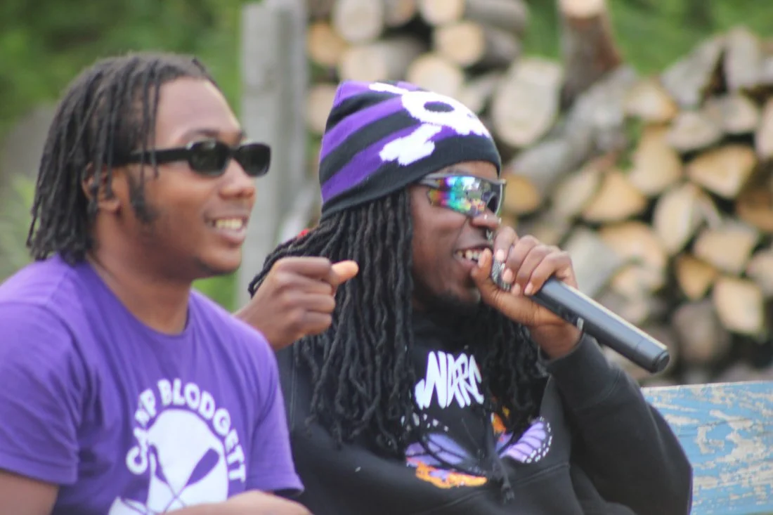 Two men sitting outdoors, wearing sunglasses and casual clothing, with one holding a microphone and speaking, and a background of stacked firewood.