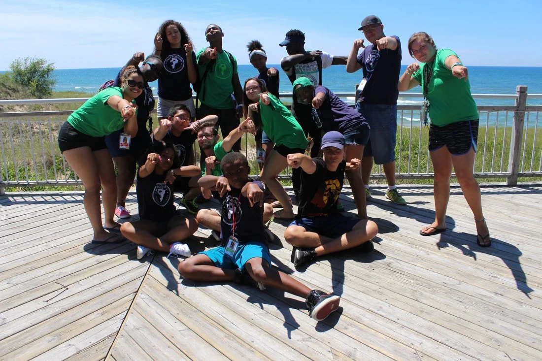 Group of young people posing on a wooden deck near the ocean, many making flexing poses.