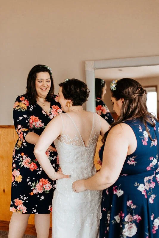 Two women in floral dresses helping a bride in a white lace wedding gown with thin straps, as they prepare for her wedding.