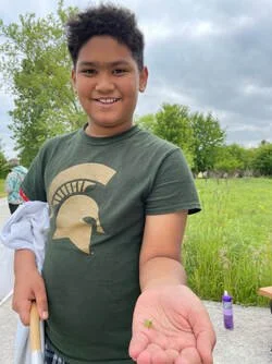 A smiling young boy holding a small green plant in his hand outdoors in a grassy area with trees and other people in the background.