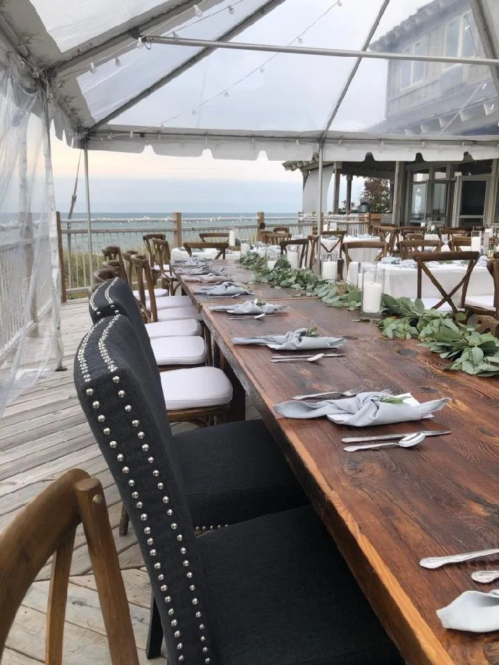 Long wooden dining table set under a clear canopy, decorated with a greenery garland and candles, overlooking the ocean with a coastal view in the background.