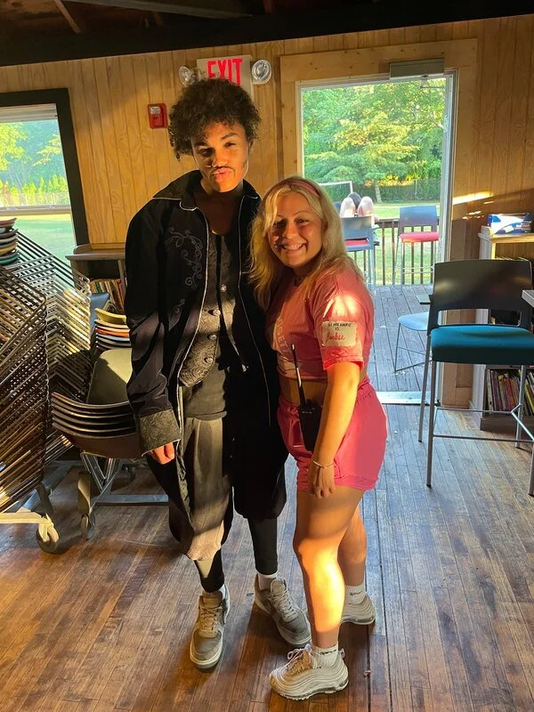 Two women standing indoors, smiling at the camera. One woman has curly dark hair and a dark jacket, and the other has blonde hair, a pink outfit, and white sneakers. Behind them is a wooden wall, with chairs, a window, and a deck outdoor area visible