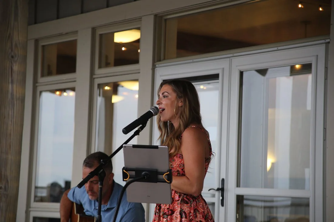 A woman singing into a microphone with a tablet attached to a stand, indoors with large windows in the background, accompanied by a man playing a guitar.