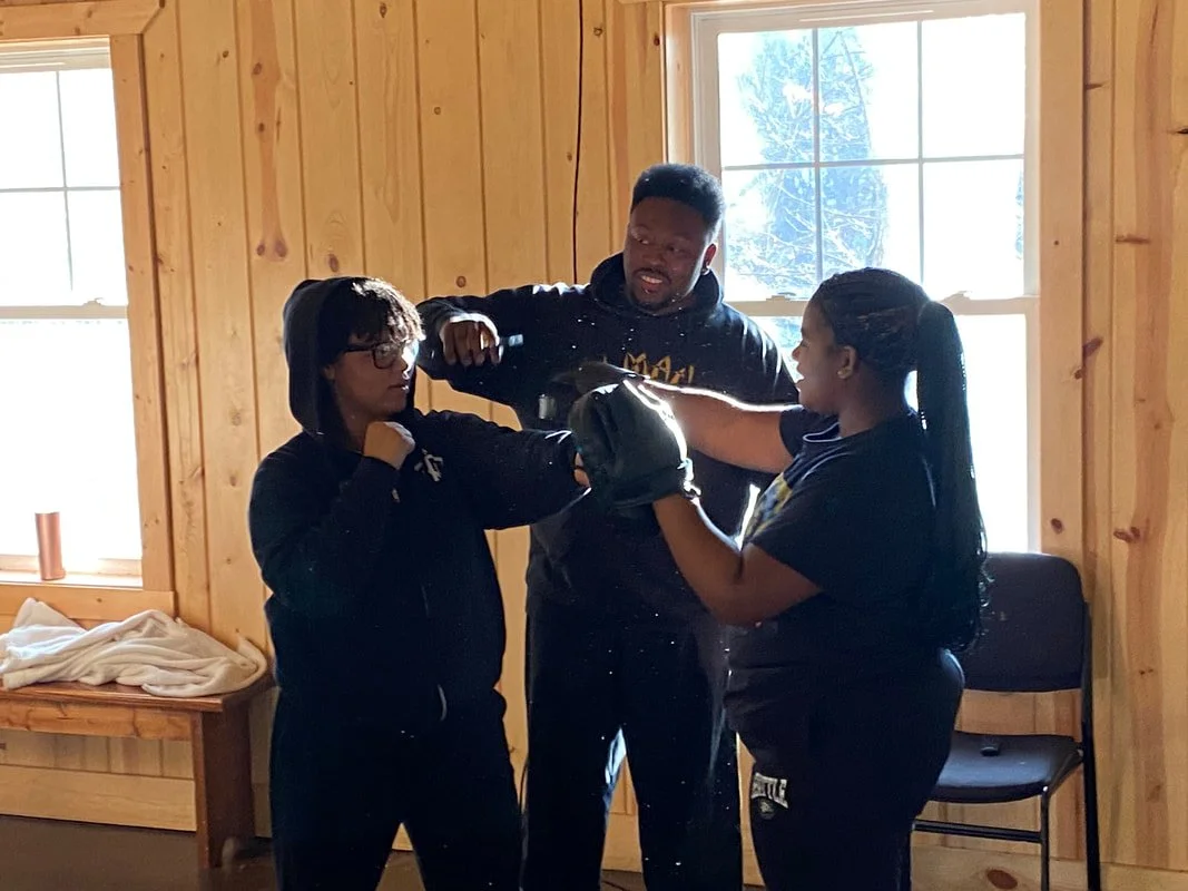 Three people practicing boxing in a wooden room by a large window, with one person holding boxing pads and the others wearing gloves.