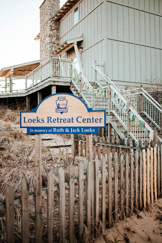 Sign for Loeks Retreat Center in memory of Ruth and Jack Loeks, with a building and stairway decorated with colorful flags in the background.