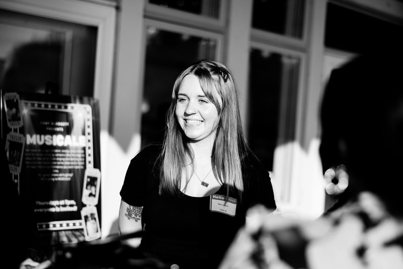 A young woman with long hair smiling and wearing a name tag, standing near a display board with the word 'MUSICAL' visible, in a well-lit indoor setting.