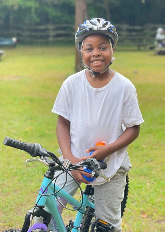 A young girl wearing a bike helmet and a white t-shirt is smiling while standing with a light blue bike in a grassy yard outdoors.