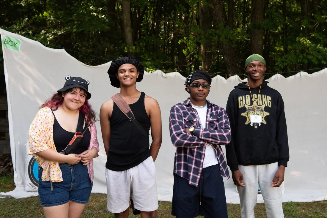 Four young people standing outdoors in front of a white sheet and trees, smiling at the camera.