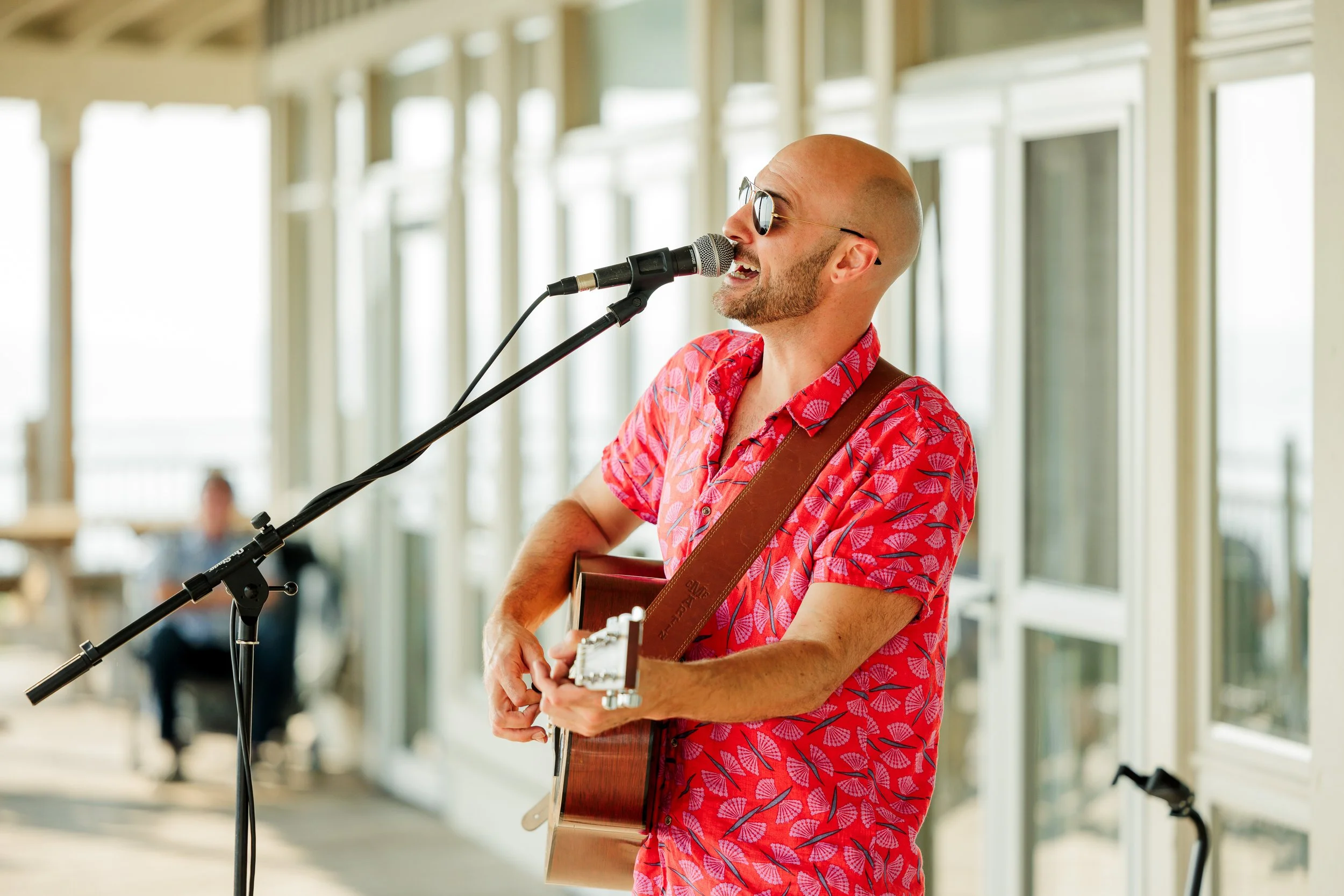 Man with bald head, sunglasses, and a red Hawaiian shirt playing guitar and singing into a microphone on a balcony.