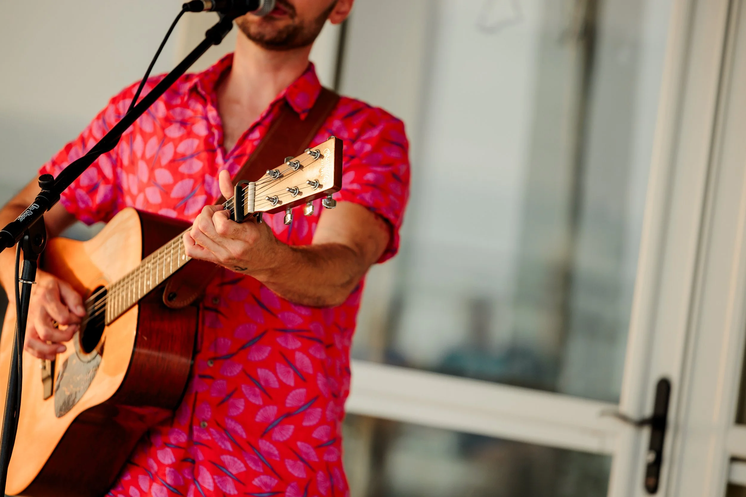 A man in a red patterned shirt playing an acoustic guitar and singing into a microphone.