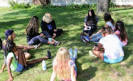 A group of children and adults sitting in a circle on a grassy lawn, having a discussion or storytime outdoors.