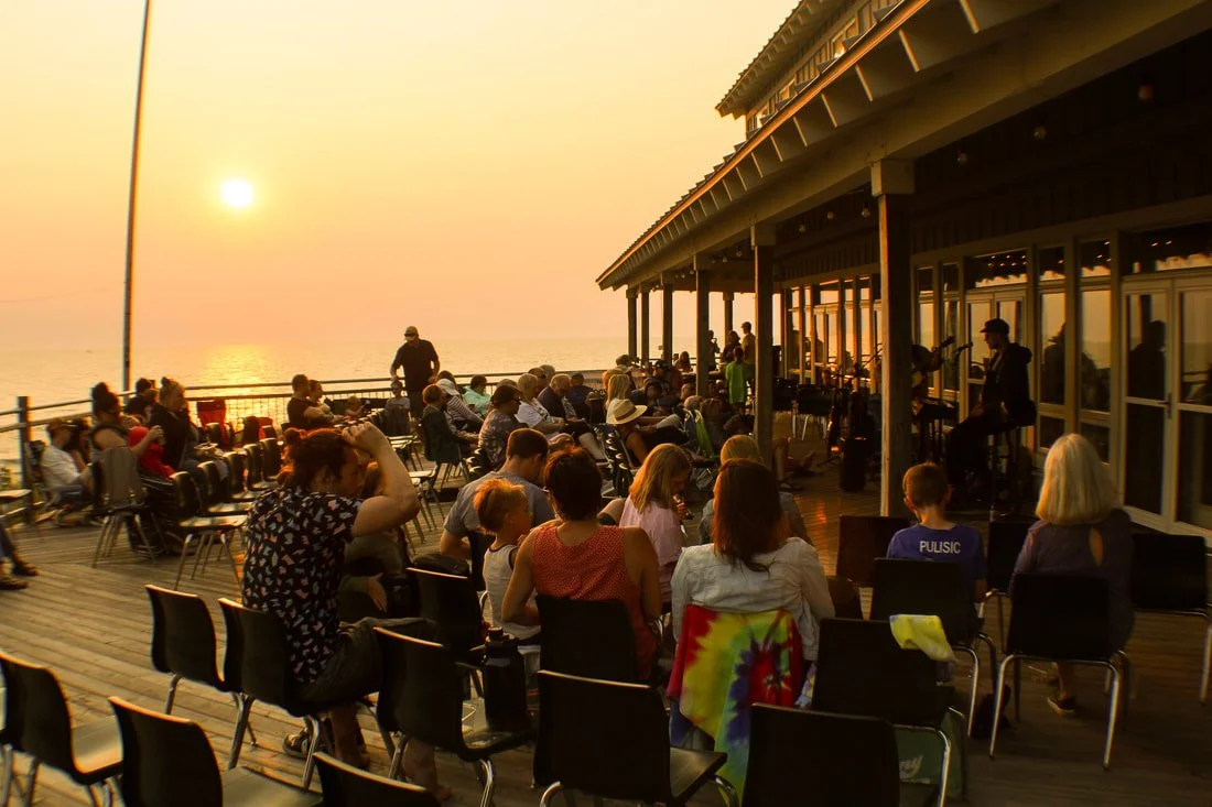 People gathered on a wooden deck watching a live music performance at sunset, near the ocean.