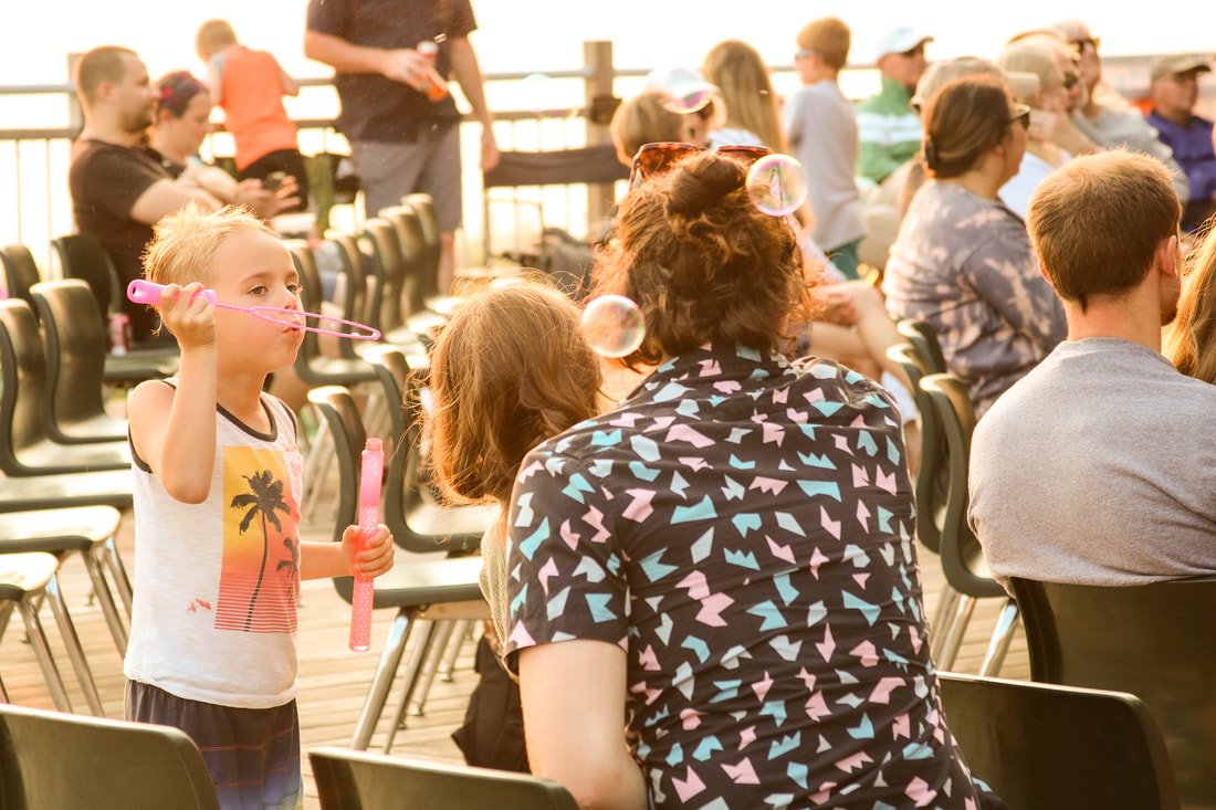 A young girl blowing bubbles with a bubble wand amidst seated adults and children on an outdoor deck.