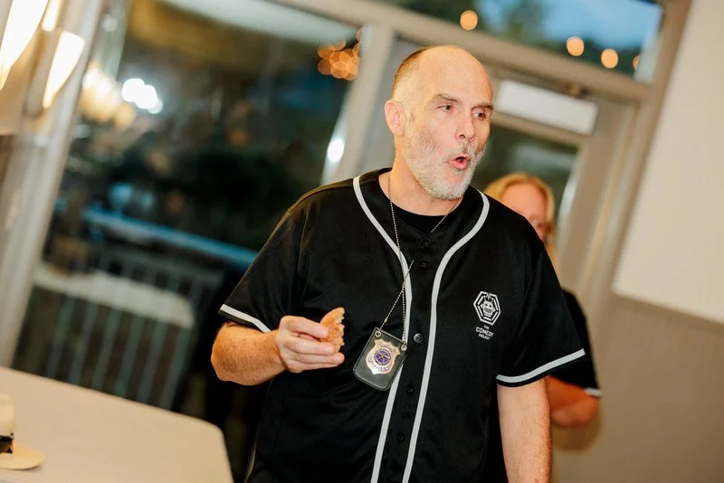 A man in a black baseball jersey with white trim holding a cookie in his right hand, wearing a badge around his neck, in a restaurant or cafe setting.