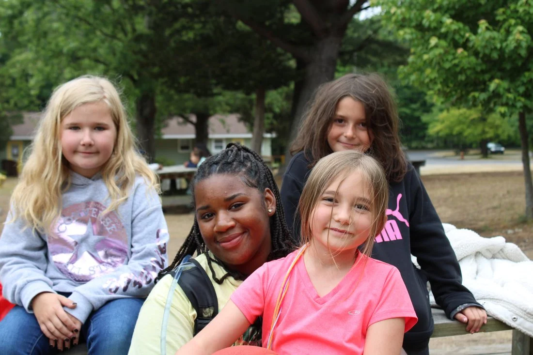 Four young girls sitting together outdoors in a park, smiling and posing for the photo, with trees and a pavilion in the background.