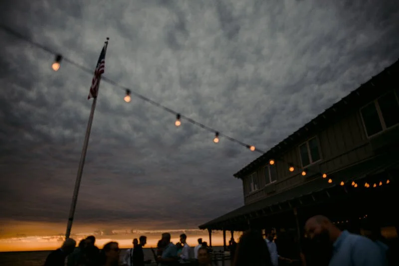 People gathered outdoors at sunset with string lights and an American flag, near a building with a balcony, on a cloudy evening.